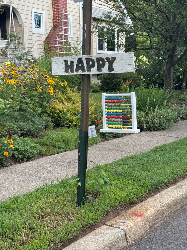 a stop sign post with a hand-made white wooden sign attached to it. the white wood sign has the word "HAPPY" hand painted in black letters. hanging from the wood sign is an abacus 🧮 ... an object commonly used to do sums. 