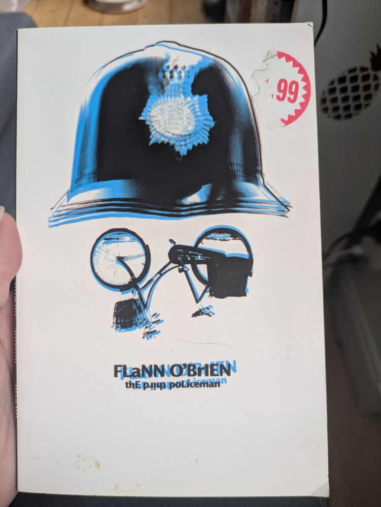 A copy of The Third Policeman by Flann O'Brien. The cover looks like a face, wearing a Policeman's cap, and an upside down bicycle in place of glasses. A price sticker is incompletely peeled off in the right corner