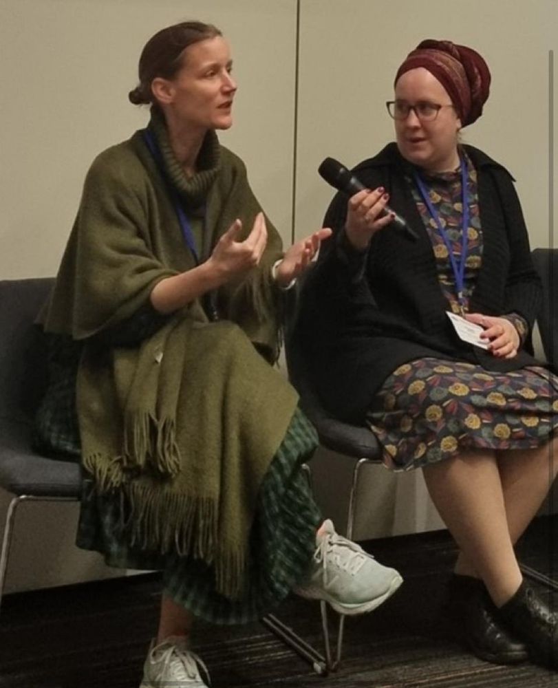 Inge, a white woman dressed in a green poncho shawl and green checked dress, speaking animatedly while Maggie, a white Jewish woman wearing a red turban-style Tichel and a blue dressed with art deco style leaves, holds a microphone toward her colleague