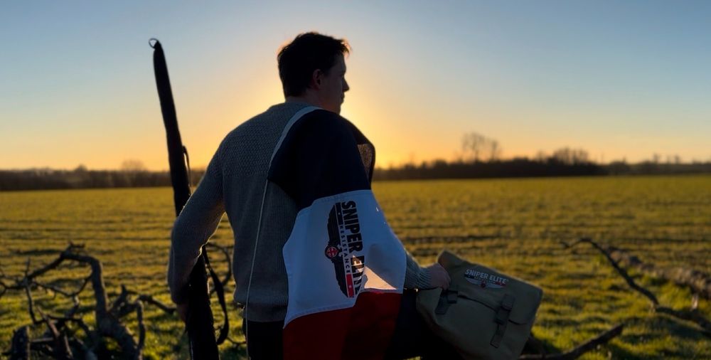 Matty stands in front of the sunrise with a French Flag and the Sniper Elite logo in the middle. A rifle and soldier's bag are also in view.