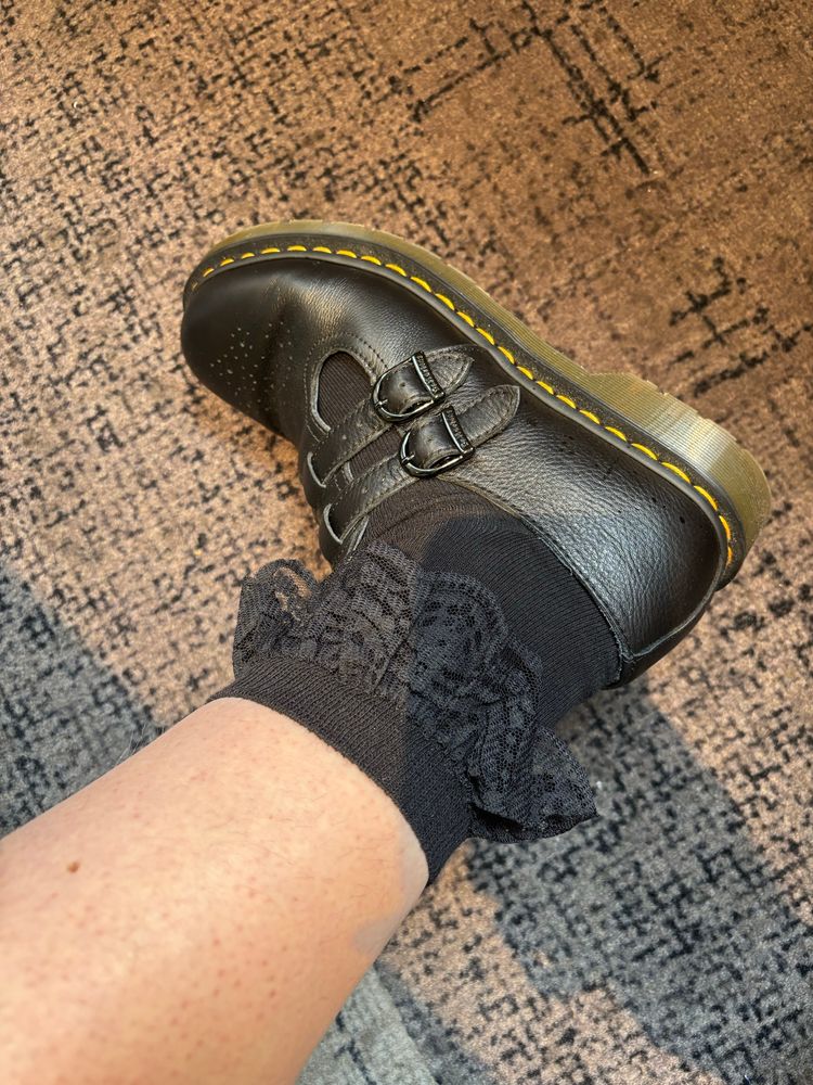 Photograph of an ankle and foot wearing black lace-topped socks and black buckle-down Mary-Jane-style shoes. 