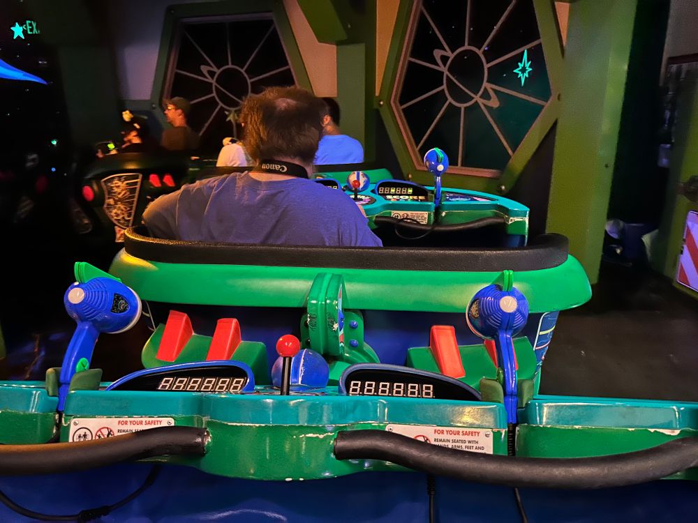 Rear view of a man in a Buzz Lightyear ride vehicle from Disneyland. The photo is taken from the right with the man sitting on the left in the vehicle in front. 