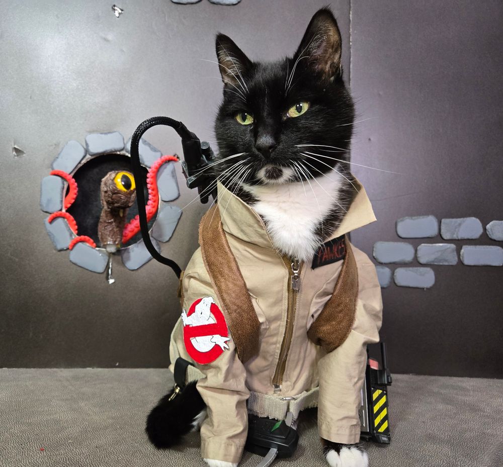 A cat in a tan Ghostbusters suit with a proton pack and ghost trap and pke meter, sitting in a sewer with a eyeball entity creeping up behind.