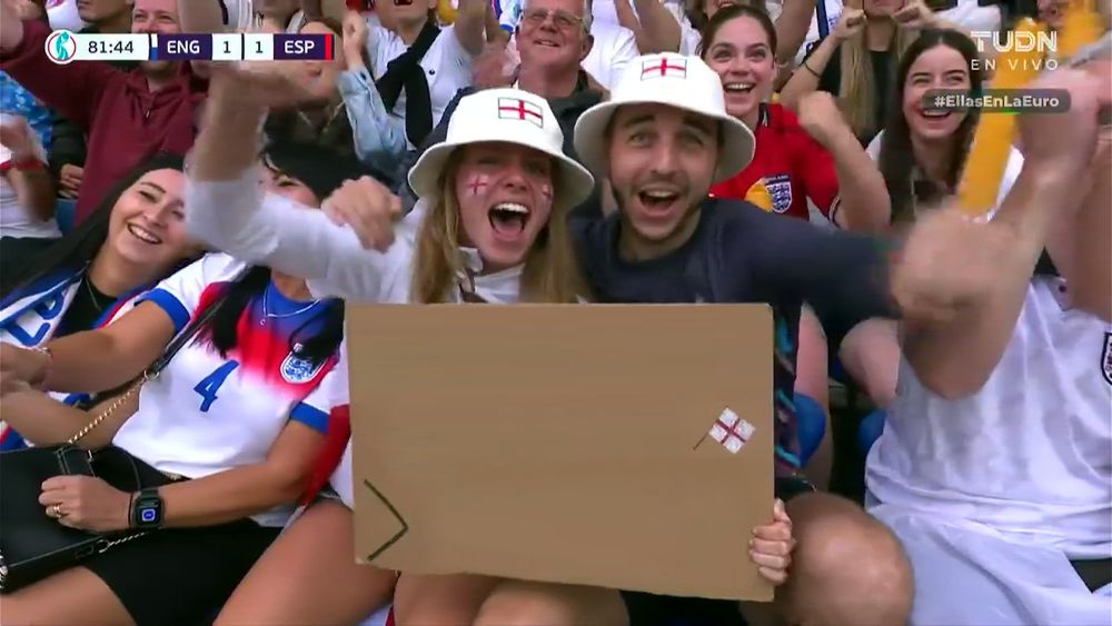 England fans holding a cardboard sign. Sign has a gap, then an England flag, a greater than symbol then another gap. The phrase "Bangers & Mash > Chorizo" has been edited out