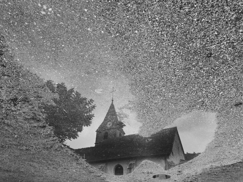 Zwart-witfoto van een kerktoren en bomen, weerspiegeld in een plas water op een grindpad. De reflectie is duidelijk zichtbaar in het water, terwijl de randen van de plas en het grind eromheen het beeld omlijsten. Het lijkt alsof je naar de lucht en de kerk kijkt, maar het is eigenlijk een spiegeling op de grond. De foto heeft een dromerige en artistieke sfeer.