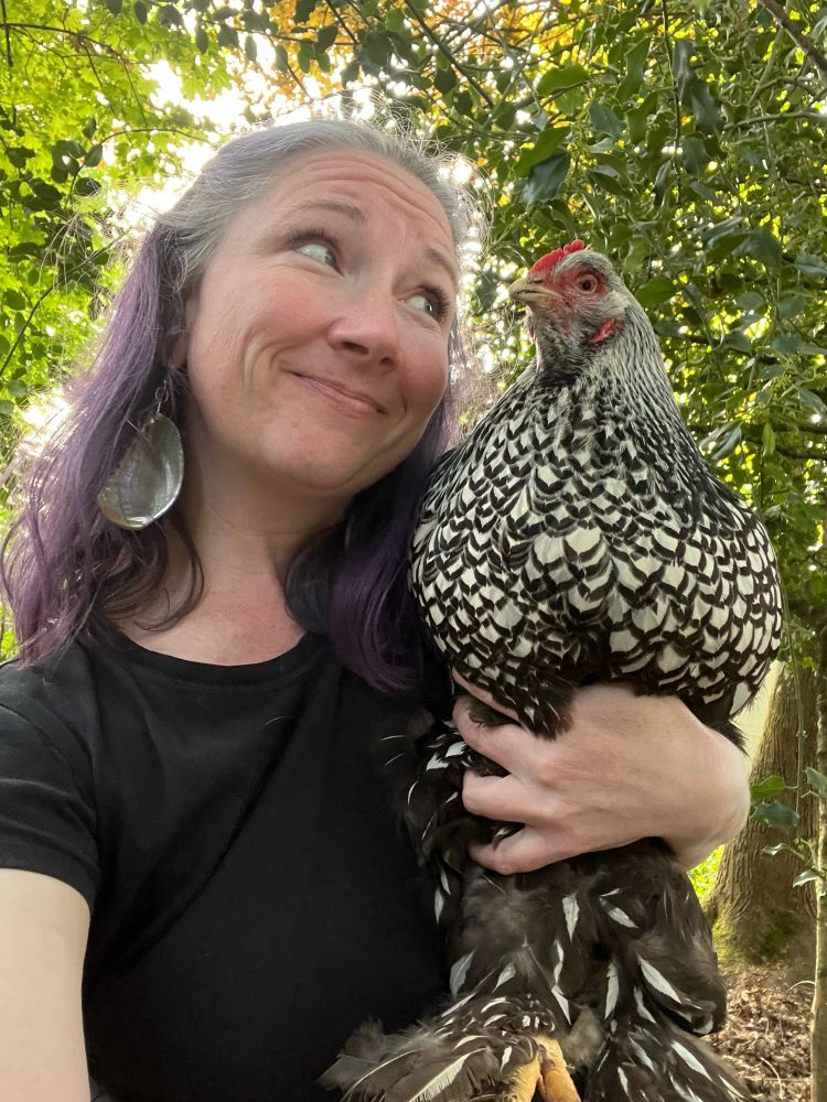 Woman with shoulder length purple hair, a black shirt, and abalone earrings. She is holding a black and white chicken at eye level, in her left arm. She is smiling at the chicken, but the chicken is not smiling back. 