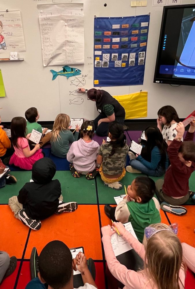Several young students sit gathered around a white board. A teacher has drawn 3 fish shapes on the board, and is adding stripes to the second fish. The first fish, with no stripes, has been “x’d” out. 