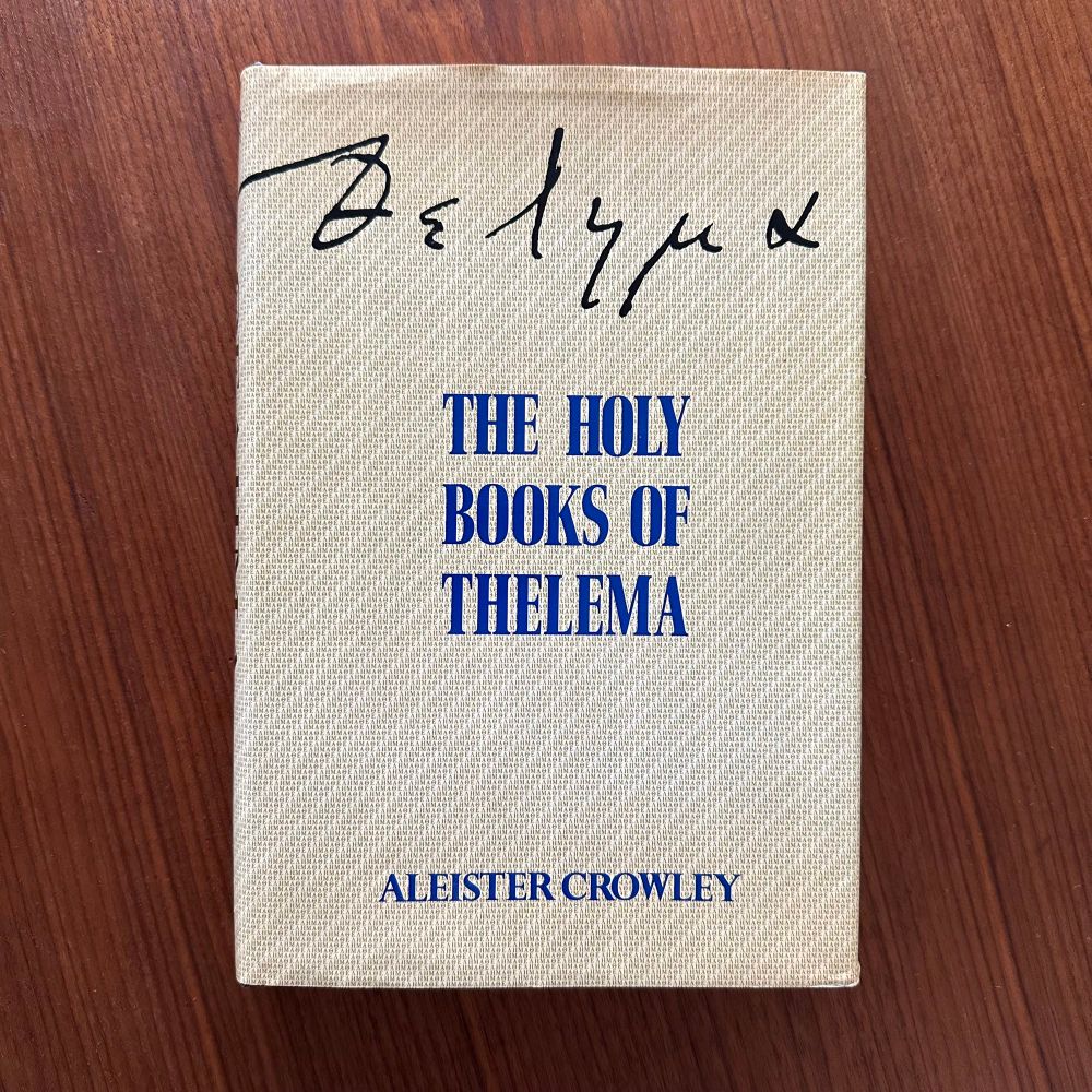 A copy of Aleister Crowley's Holy Books of Thelema, first edition. The dust cover is beige with some form of vague pattern on it, with "Thelema" written in hand in Greek at the top, the title of the book in blue in the middle and the author's name in capital letters at the bottom.