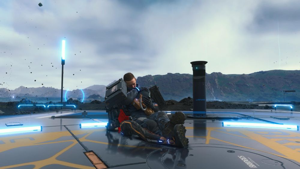 Screenshot from Death Stranding. Sam, the main character, sitting on the ground, wearing all his gear, sleeping, cradling the tank that holds the magical baby in his arm. Vista of mountains in the background and gray skies above.