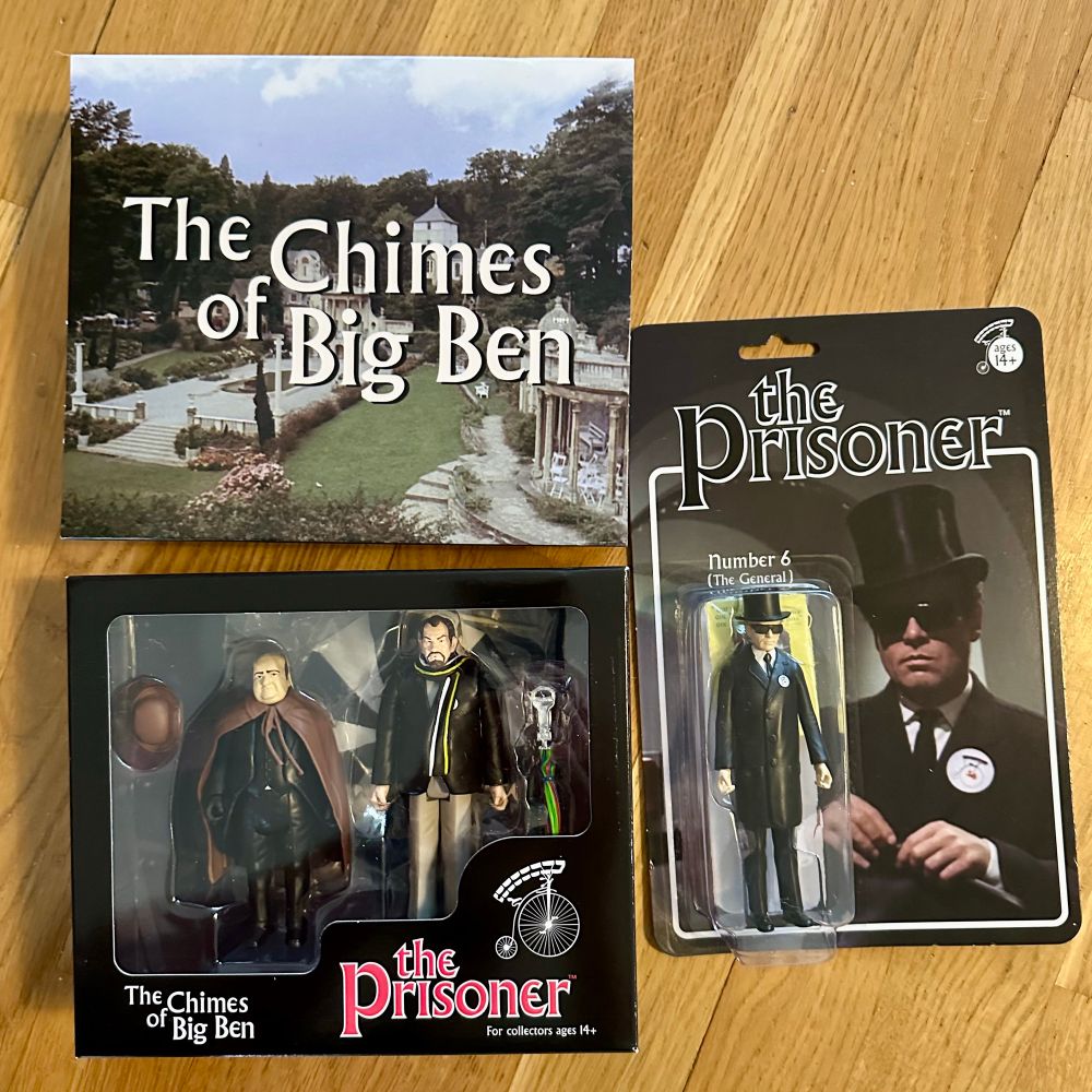 Two boxes of The Prisoner figurines. One box contains two figurines of the Butler and No 2, with its cover with the text "Chimes of Big Ben" on it lying next to it. The second box contains No 6 dressed as The General in a big top hat.