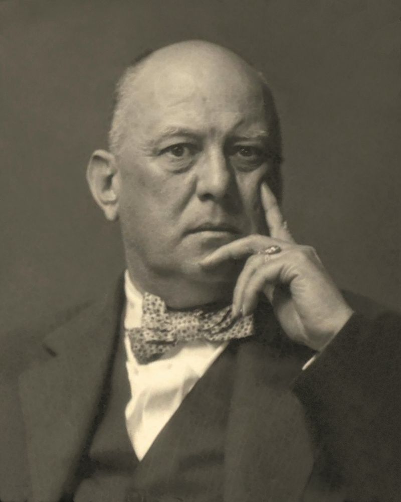 Black and white photograph of Aleister Crowley in 1925, looking into the camera, his hand against his cheek, wearing a suit and a bowtie.