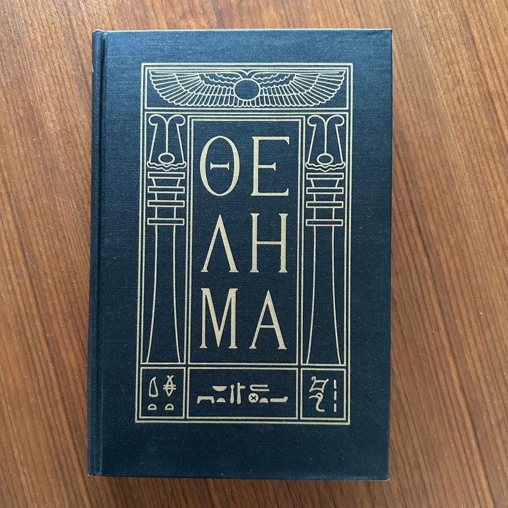 The book without the dust cover. Black, with "THELEMA" printed in capital letters, set within an Egyptian styled frame. 