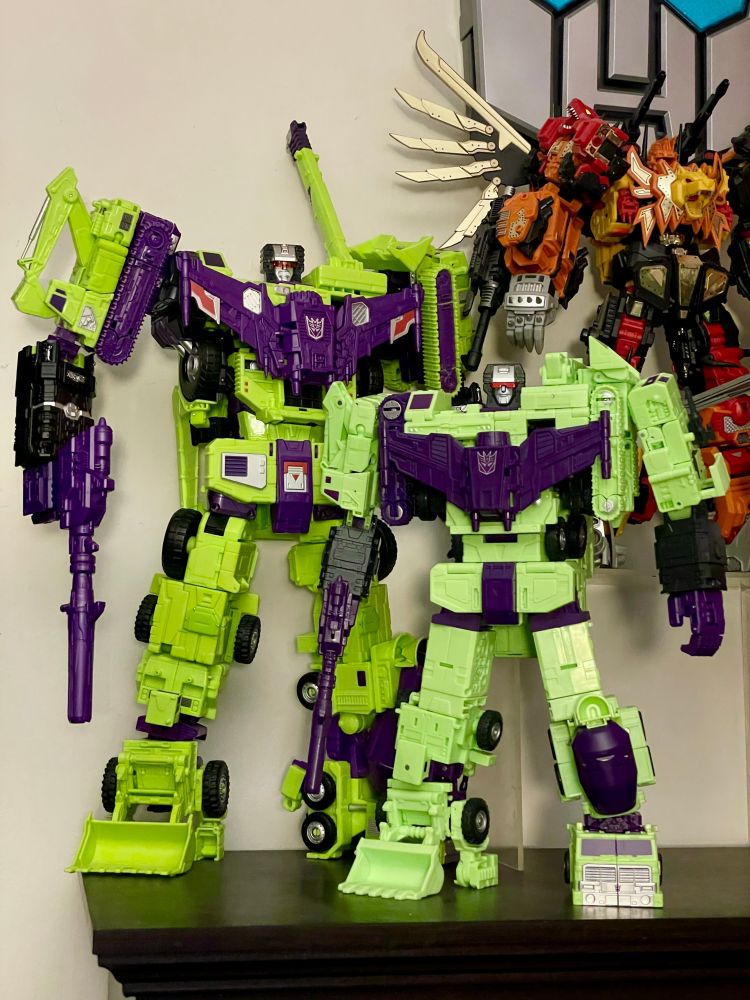 Photo comparison of Unite Warriors Devastator (left) with DNA Design upgrades that lengthen the forearms and extend the shoulders out (among a few other things), and the new Studio Series 86 Devastator (right). With a little Mastermind Creations Feral Rex (with reprolabels) hanging out in the background.