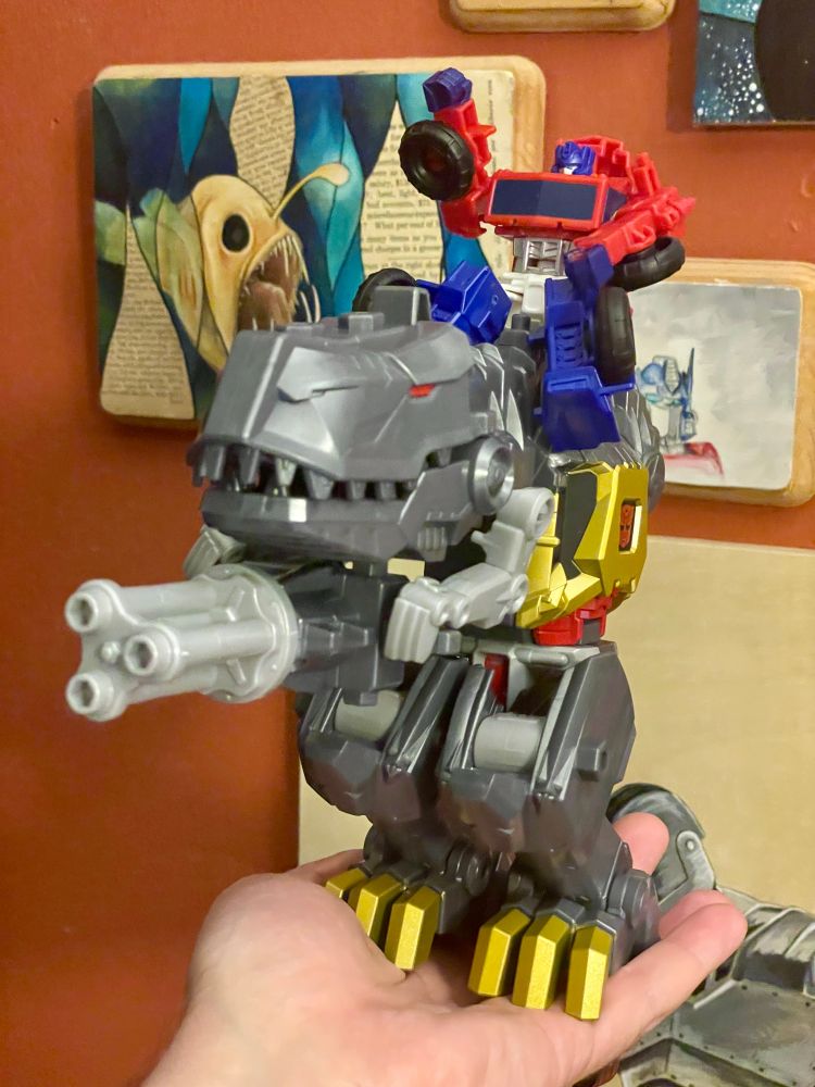 Transformers Cyberworld Grimlock in dino mode, holding his gatling gun tail in his tiny rex arms like some kind of goofy Warhammer mount, with Cyberworld Optimus Prime riding on his back. This thing is so ridiculous, I love it.