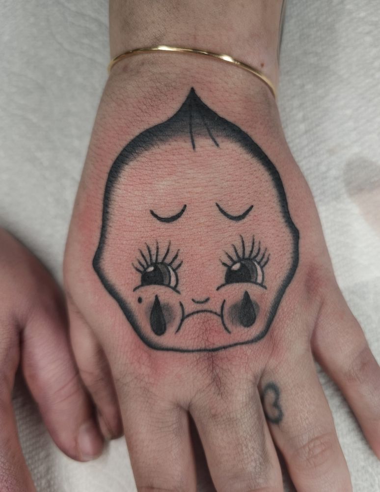 Hand thats been tattooed, now shows a kewpie baby face that is sad and has 2 tears on either cheeks. Is frowning, eyebrows turned down, and has long eyelashes. Done in black and grey and has a vintage look