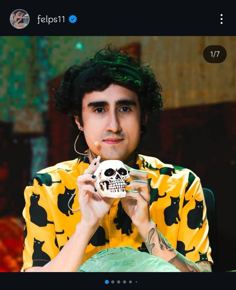 foto do felps segurando uma caveira (de mentira), ele está sorrindo levemente com uma blusa amarela com estampa de gatos pretos
