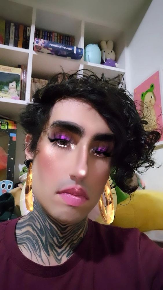 Foto do Felps na frente de uma estante branca com livros e bichos de pelúcia. Ele está vestindo uma blusa vermelha escuro e usando um filtro do Instagram que deixa ele com o rosto harmonizado e maquiagem.