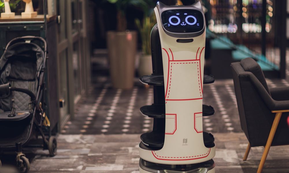 A robot waiter with a smiling cat face, painted like it is wearing overalls. It has a cylindrical body about four feet tall, with three levels of trays to place dishes on. Pudu's Bellabot. 