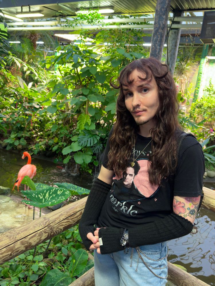 An androgynous looking twink with long wavy hair and colourful tattoos poses in front of a flamingo whilst wearing an "Edward" from Twilight shirt (unironically).