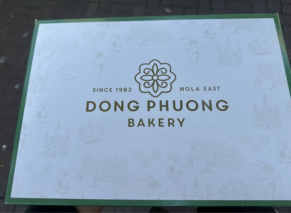 Image of the top of a king cake box that is white with a green border. Printed in gold are an icon of a moon cake and the words “Since 1982, New Orleans East, Dong Phuong Bakery”
