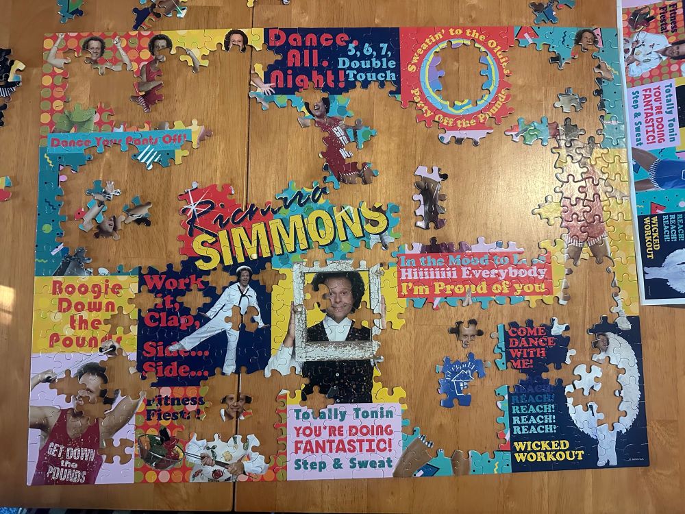 A partially put together jigsaw puzzle on a wooden table. The puzzle depicts multiple pictures of Richard Simmons.