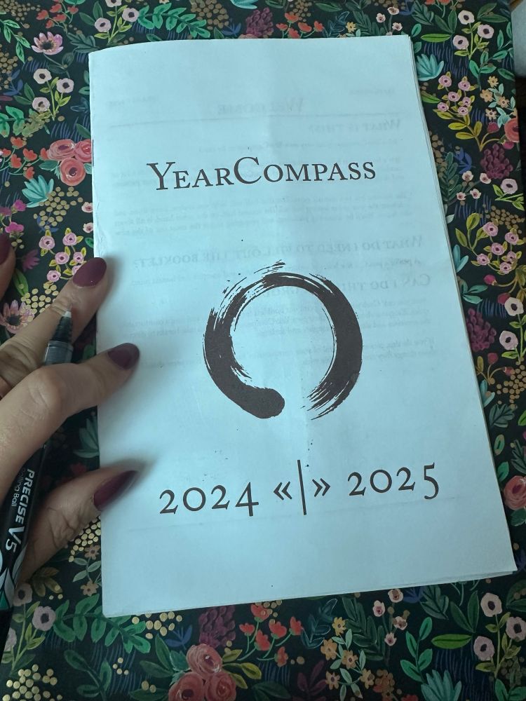 Photo of front cover of printed pamphlet that says “YearCompass” with a circle paint stroke in the middle and “2024 <<|>> 2025” with my left hand and pen on the side. Pamphlet is on a pretty dark floral clipboard 