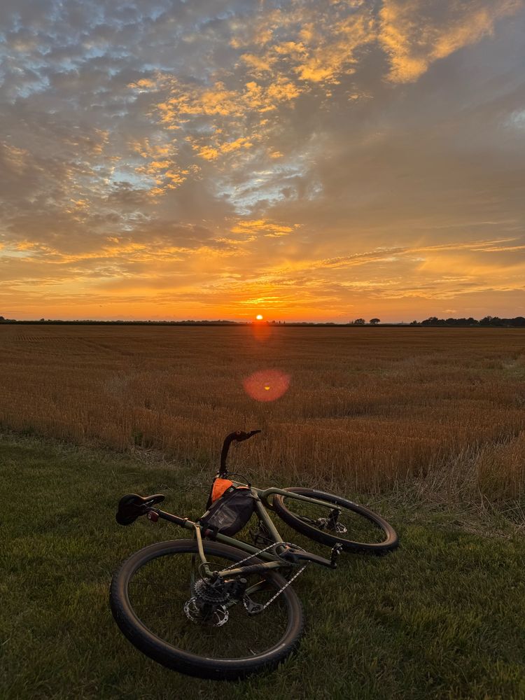 A gravel bike on the ground next to a cut wheat field. The sun is setting and casting a reflection on the clouds. 