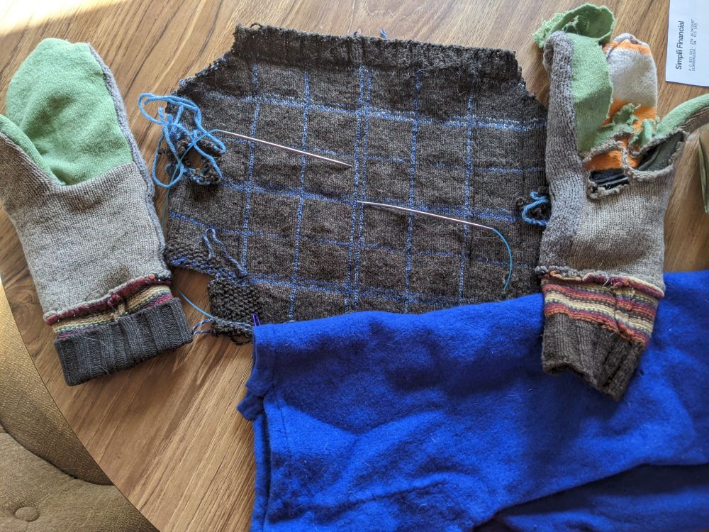 A pair of mittens, with the palm made of two colours of contrasting fabric.  One of the mittens looks like it has been attacked by a belt-sander.  Next to the mittens is the tartan knit from the previous bluup and a similar sized blue piece of felt that looks like it was stored in a crumpled up ball