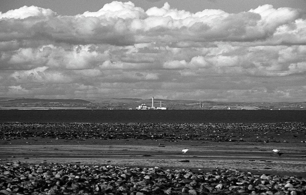 A black and white photo looking across to Wales from Dunster Beach on a clear day. There are a couple of waders on the beach and a factory in the distance in South Wales. 