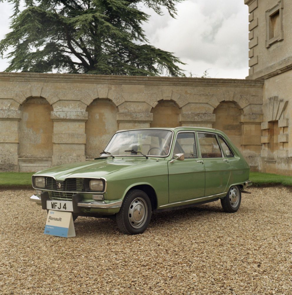 A colour photo of an old green Renault parked on gravel in front of a sandstone wall.
