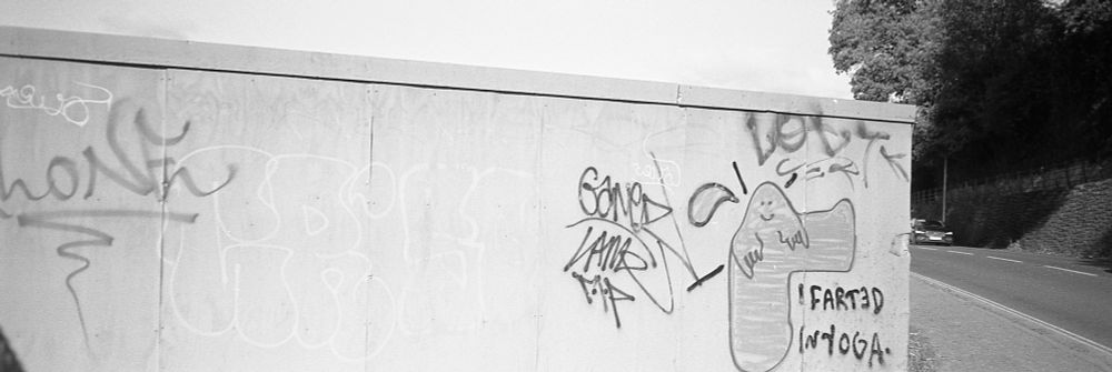 A black and white photo of a graffiti strewn hoarding which shows a figure with a raised leg stating 'I farted in yoga'.