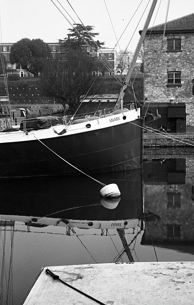 A black and white photo of the bow of a large boat called Snark with its reflection in the river and buildings behind.