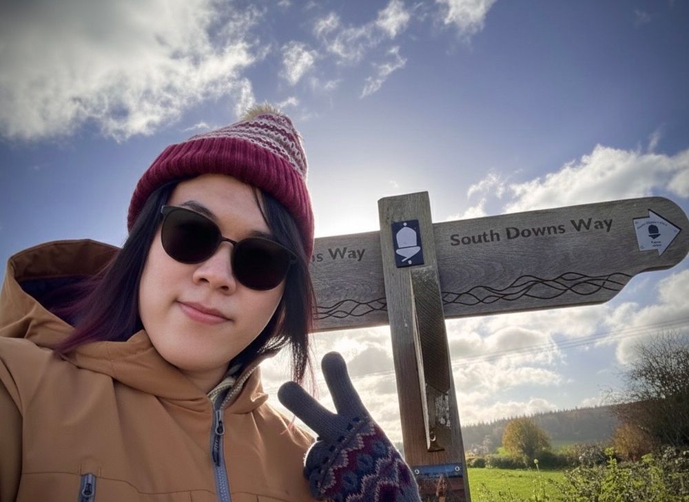 A low-angle selfie shows a person wearing a maroon knit beanie, sunglasses, and a tan jacket, holding up a v sign with a gloved hand. They are standing next to a wooden trail marker for the "South Downs Way," which has an acorn symbol and an arrow pointing right, all set against a bright, cloudy blue sky.