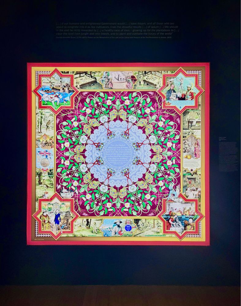 A photograph of a large, framed square artwork hanging on a dark wall, illuminated by gallery lighting. The artwork is designed to resemble a luxury silk scarf with an intricate, symmetrical pattern.
The center features a light blue, lace-like medallion containing text, surrounded by a deep magenta field filled with stylized green vines and white opium poppies. The wide, golden-yellow border contains detailed illustrated vignettes—some inside eight-pointed stars and others in rectangular panels. these illustrations depict historical scenes related to the British Empire, the tea trade, and the Opium Wars, showing figures in colonial military uniforms, ships, and laborers. Faint white text explaining the context is printed on the wall above the frame.