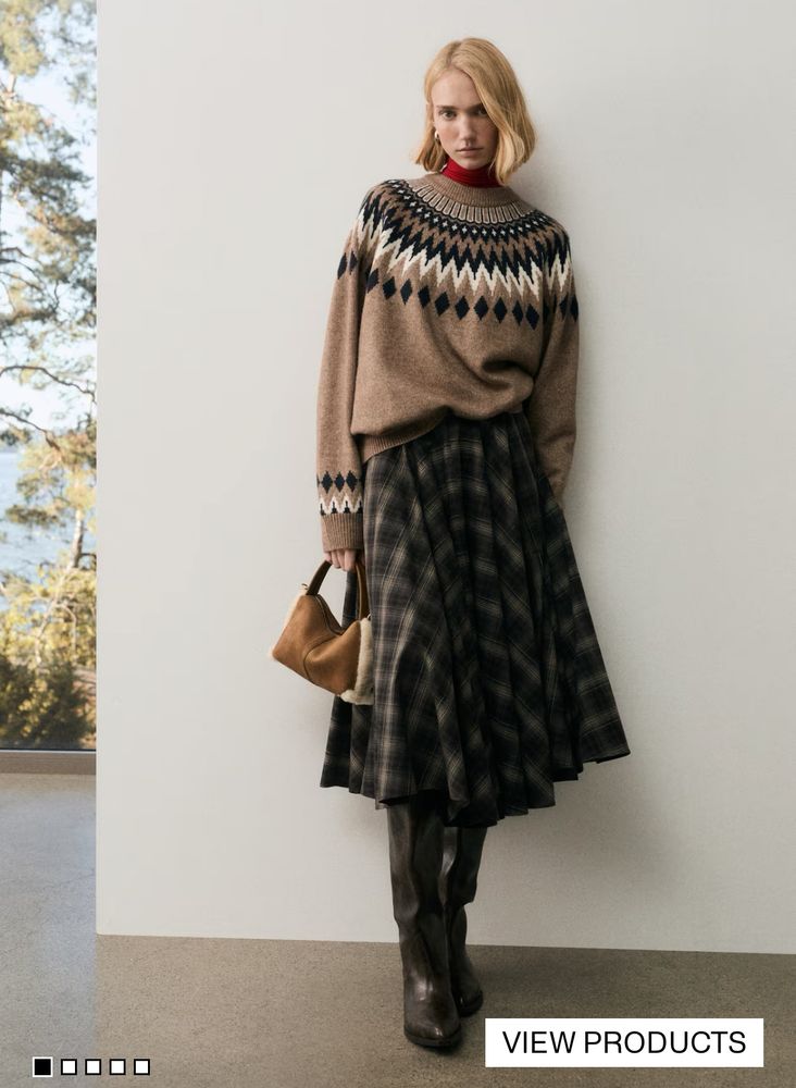 A fashion model stands indoors, wearing a light brown oversized Fair Isle sweater over a red turtleneck, paired with a dark, flowy plaid midi skirt and tall dark leather boots. She holds a small brown shearling bag.
