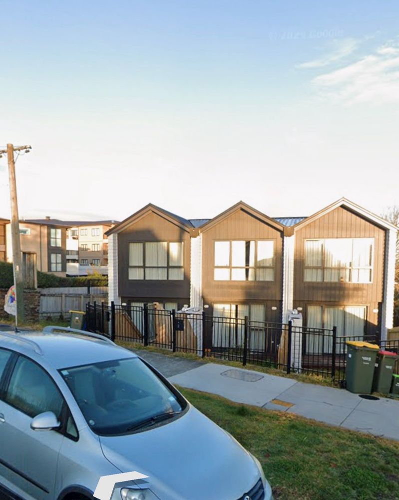 A street-level view of three new build houses featuring dark vertical siding, white trim, and pointed gabled roofs. A black metal fence runs along the front of the properties, separating the small yards from the sidewalk where green and yellow wheelie bins are placed. The sky is blue with light clouds.
