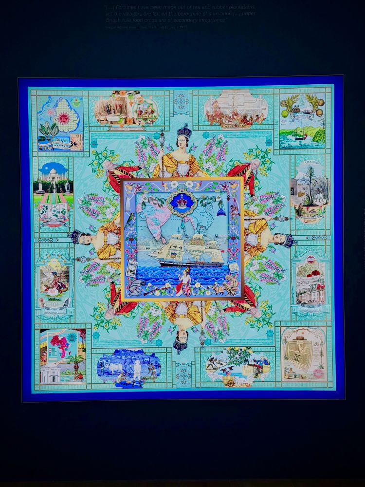 A framed, square artwork with a complex, symmetrical design resembling a high-end silk scarf, displayed against a dark blue wall. The artwork features a light turquoise background with a central square illustration of a sailing ship, a world map, and a crown. Radiating outward from the center are four figures: two depictions of Queen Victoria holding a scepter and two men in red military uniforms. The outer border is lined with detailed rectangular vignettes depicting scenes from the British Empire, including maps of India and Africa, the Taj Mahal, and illustrations of colonial labor and plantations. Above the artwork, faint text printed on the wall reads, "Fortunes have been made out of tea and rubber plantations, yet the villagers are left on the borderline of starvation [...] under British rule food crops are of secondary importance."