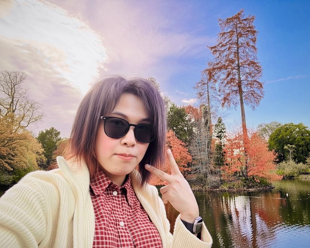 A person with purple-grey hair and sunglasses takes a selfie, holding up a V sign. They are wearing a cream cardigan over a red checkered shirt, standing in front of a pond with trees displaying bright red and orange autumn foliage in the background.