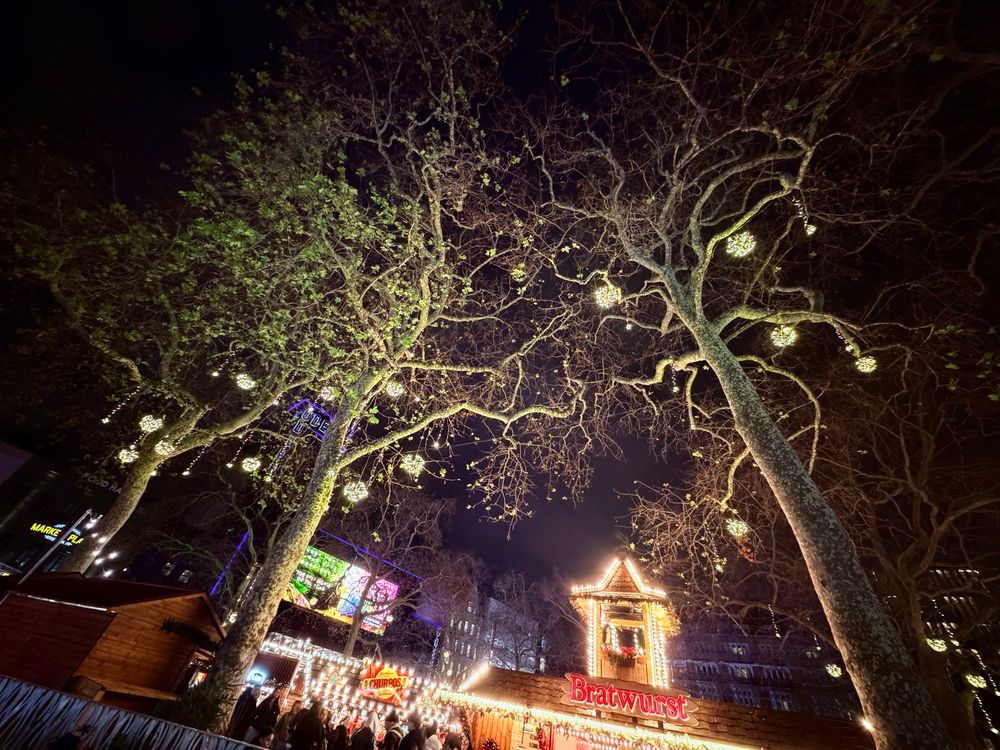 A nighttime wide-angle shot looking up through the sprawling, bare branches of large trees at a festive outdoor market. Glowing, spherical light ornaments are scattered throughout the branches against a dark sky. At the bottom of the frame, wooden market stalls are illuminated with warm lights. Colorful neon signs and the silhouettes of a crowd are visible in the background.