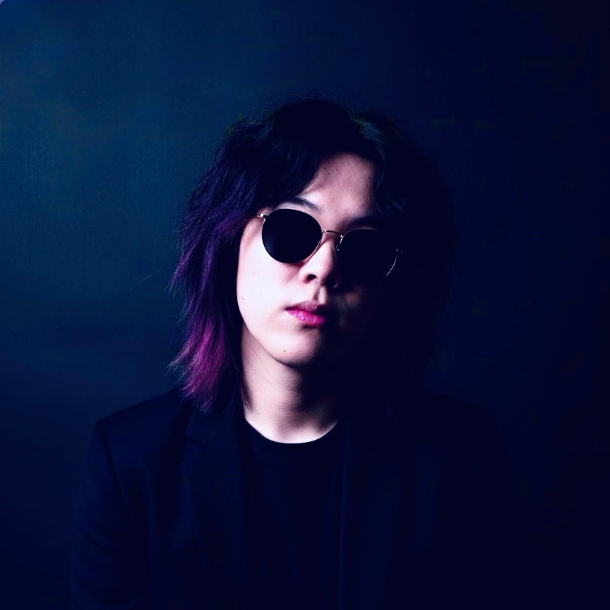 A serious-looking, androgynous person with shoulder-length, dark, textured hair that has subtle purple highlights, gazes directly into the camera.