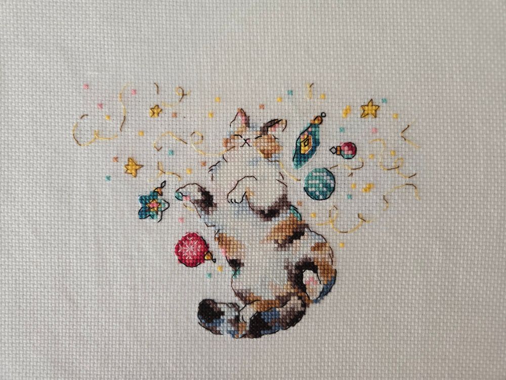 a cross stitch of a multicolored cat lying belly up with its eyes closed amidst Christmas tree decorations and confetti.