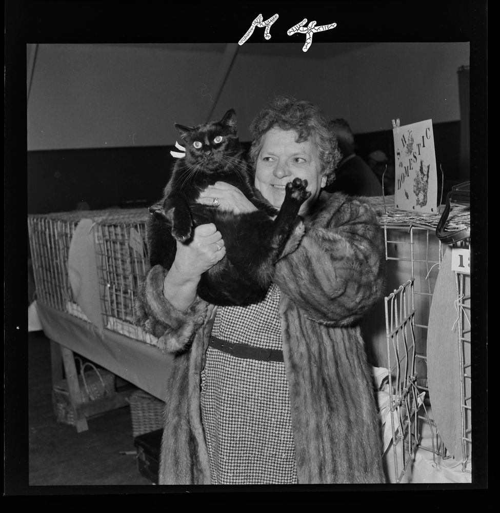 A black and white photo of a woman in a fur coat, holding an alarmed looking black cat.