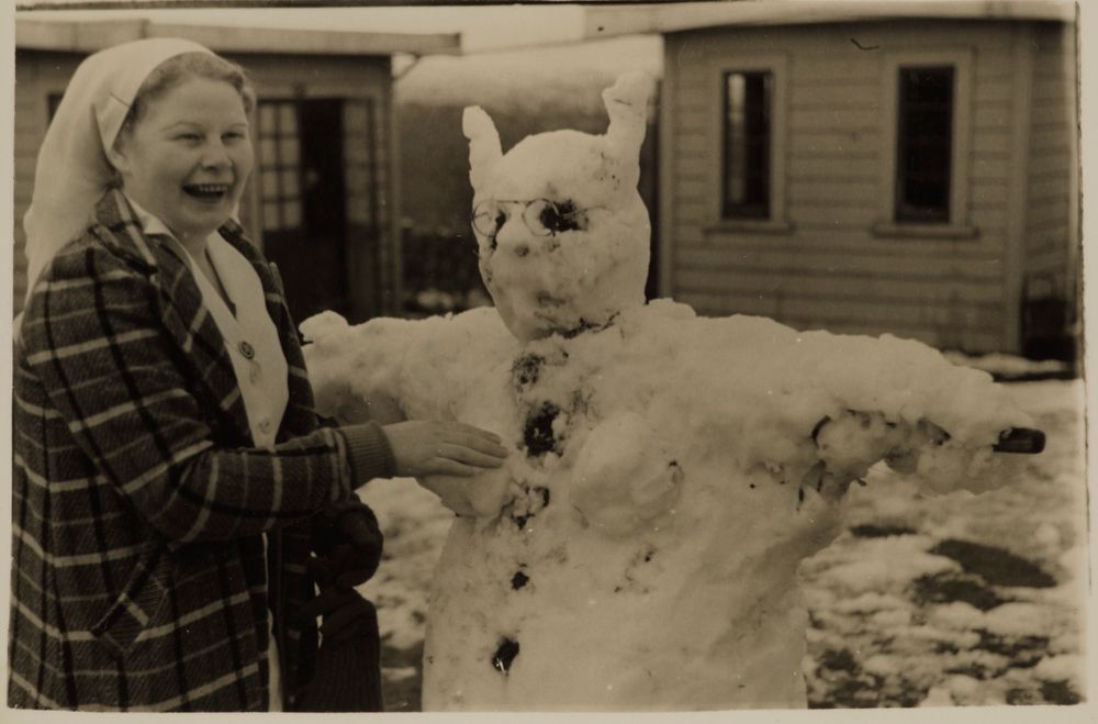 A laughing nun in an overcoat attaching snow breasts to a snow woman.