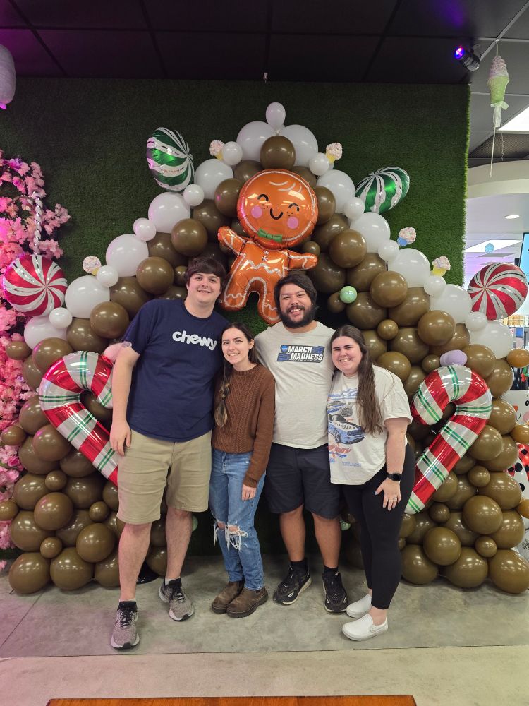 Four friends in front of a gingerbread house style balloon arch