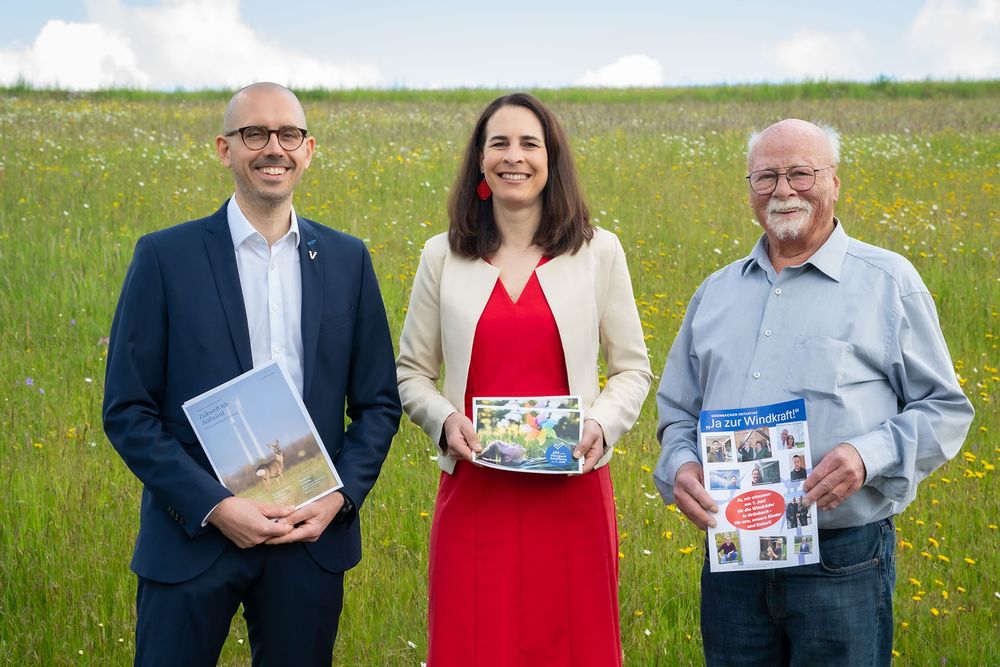 Gruppenbild mit Projektleiter Philipp Stöger, Geschäftsführerin Lisa Csenar und dem Sprecher der Bürgerinitiative Pro Windkraft Grünbach, Franz Steinmaßl. Alle drei halten Flugblätter für die Windkraft in den Händen und stehen vor einer grünen Blumenwiese,