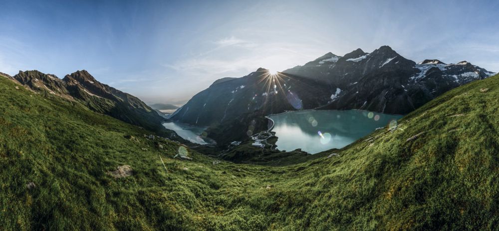 Grüne Almwiese mit Blick auf einen Stausee mit Staumauer im Licht der aufgehenden Sonne über einem Bergkamm