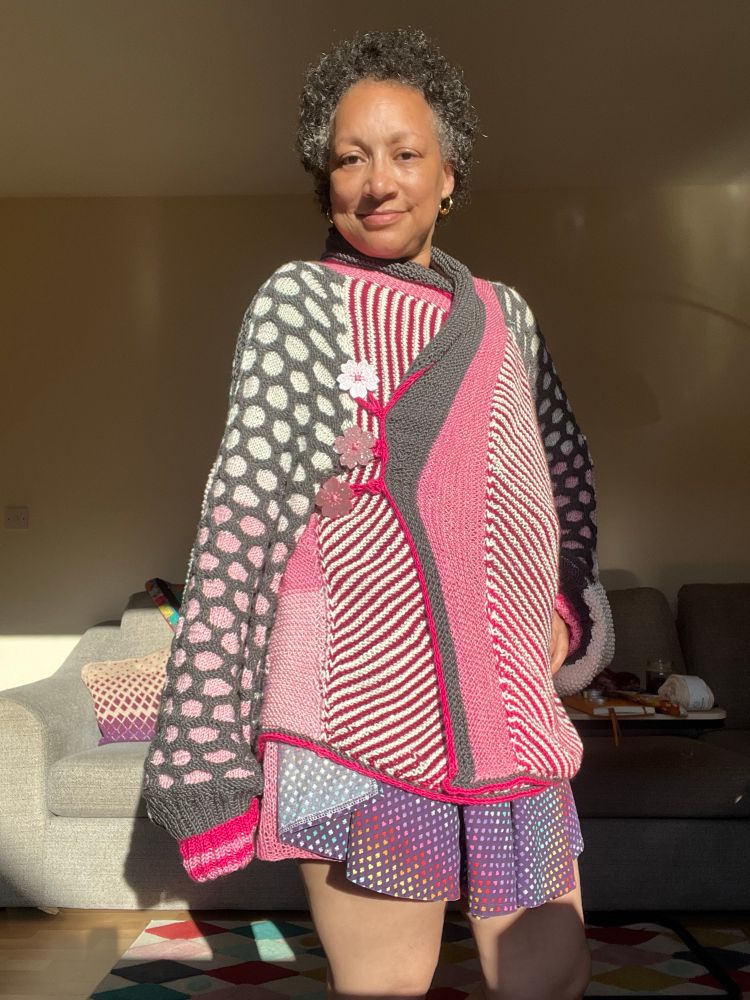 A light skinned Black woman wears a knit jacket. The arms are honey comb knit with grey lines and graduate from white to pink. The cuffs are hot pink with pink stripes. The front of the jacket is fuchsia and cream stripes. The collar is pink & grey and the whole thing is edged in hot pink. There are three giant glittery pink buttons in the shape of Sakura. 
