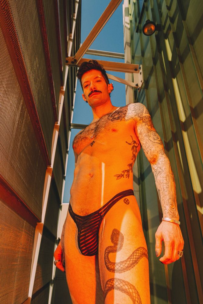 Low shot of RJ standing in a strong pose, looking down at the camera, wearing nothing but a mesh thong that puts it all on display 