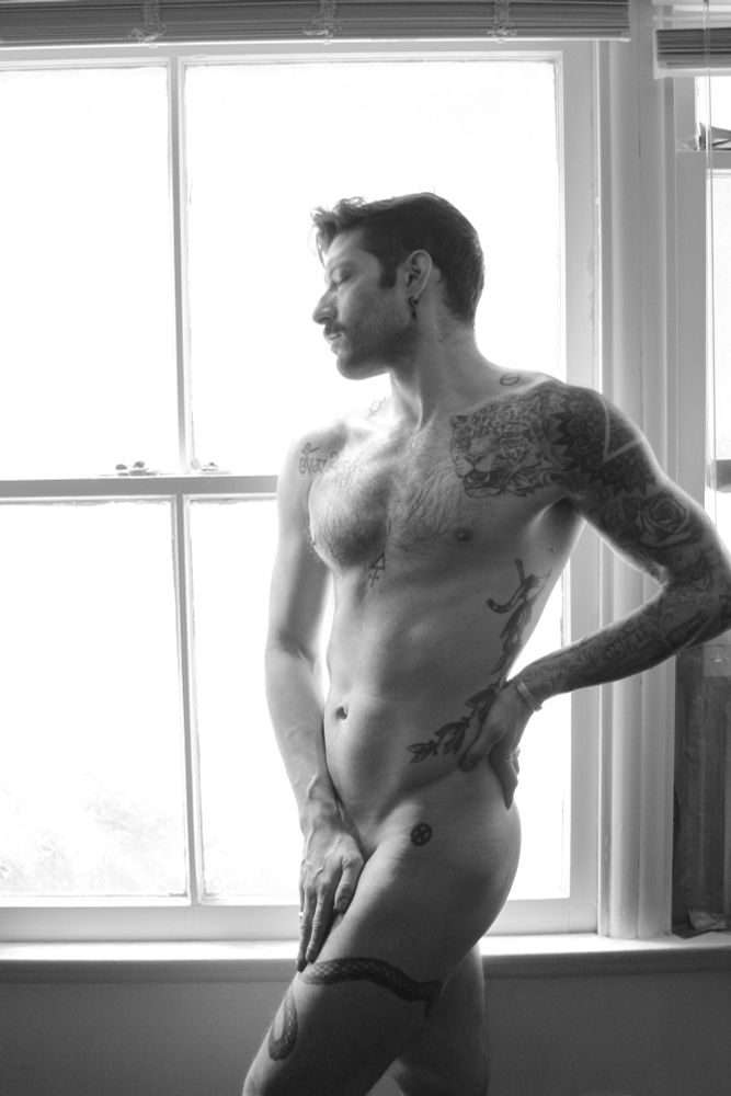 A nude RJ standing next to an open window posing with one hand on his hip and the other covering his junk. 