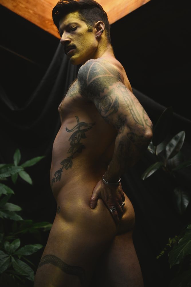 Photo of a nude RJ lit from above and covered in splashes of gold paint while groping his butt